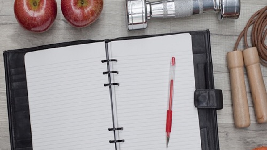 A fitness journal on a table next to 2 apples a dumbbell a jumping rope and a pair of shoe.