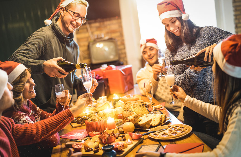 Friends group with Santa hats celebrating Christmas with champagne and sweets food at home dinner - Winter holidays concept with people enjoying time and having fun eating together. All the good food and sweets causes holiday weight gain.