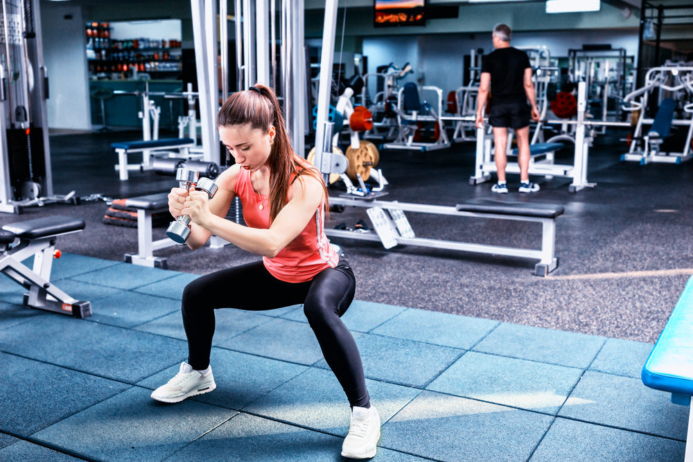 Woman doing squat and holding dumbbells in gym. Suitcase Squat is one of the exercises for functional fitness.