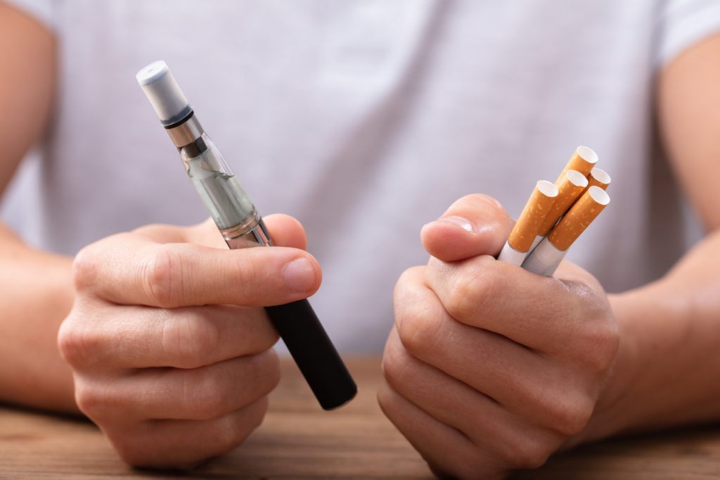 Man Holding Vape And Tobacco Cigarette Over Desk. What do you think? Is vaping bad for you?