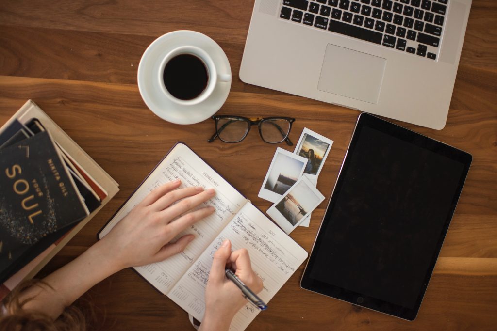 A woman writing on a notebook with her laptop, cup of coffee, reading glass, a tablet, and couple of books on a table figuring out what is life's purpose.