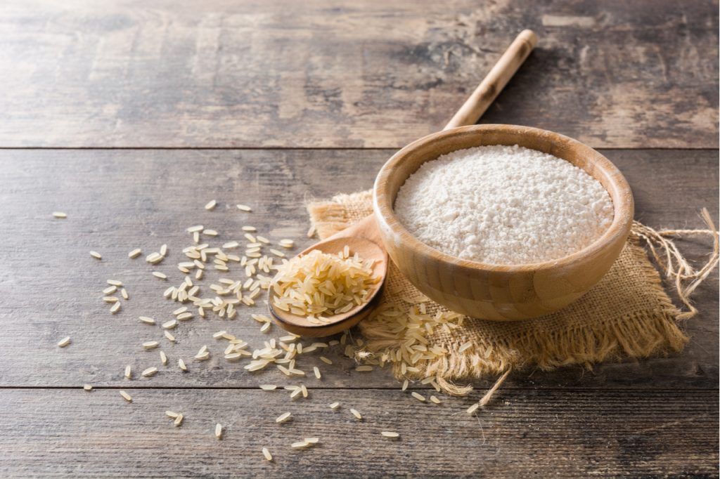 Brown rice powder in a wooden bowl, beside a spoonful of uncooked brown rice.