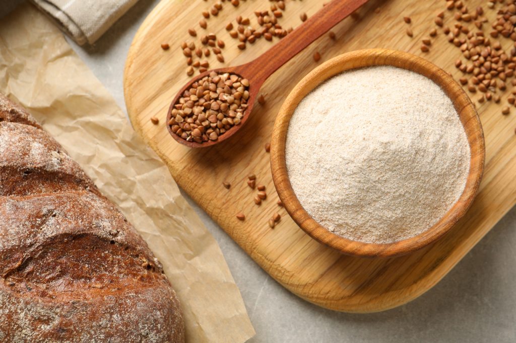Buckwheat flour in wooden bowl next to a spoonful of buckwheat on a grey table.
