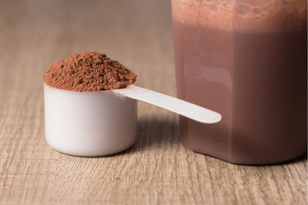 Chocolate protein powder in a measuring scoop next to a shaker bottle filled with protein shake.