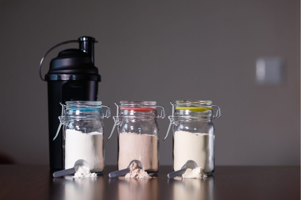 Three different protein powders in glass jars next to a shaker bottle.