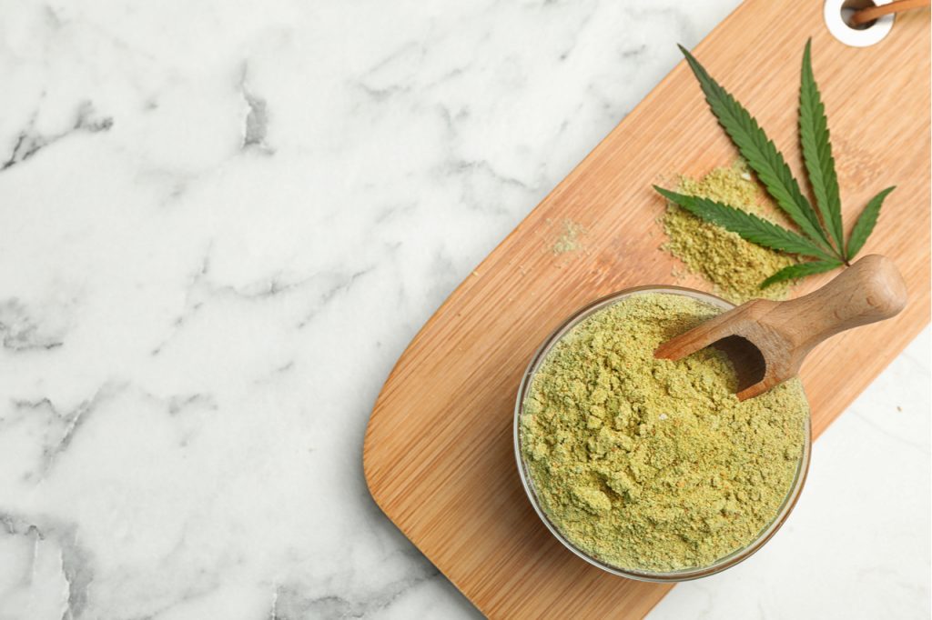 Hemp protein powder in a bowl with a leaf of cannabis on a wooden board.