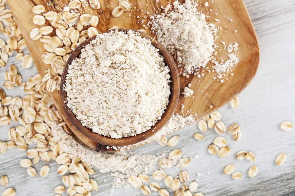 Oat wholegrain flour in bowl with oat flakes scattered on wooden table.