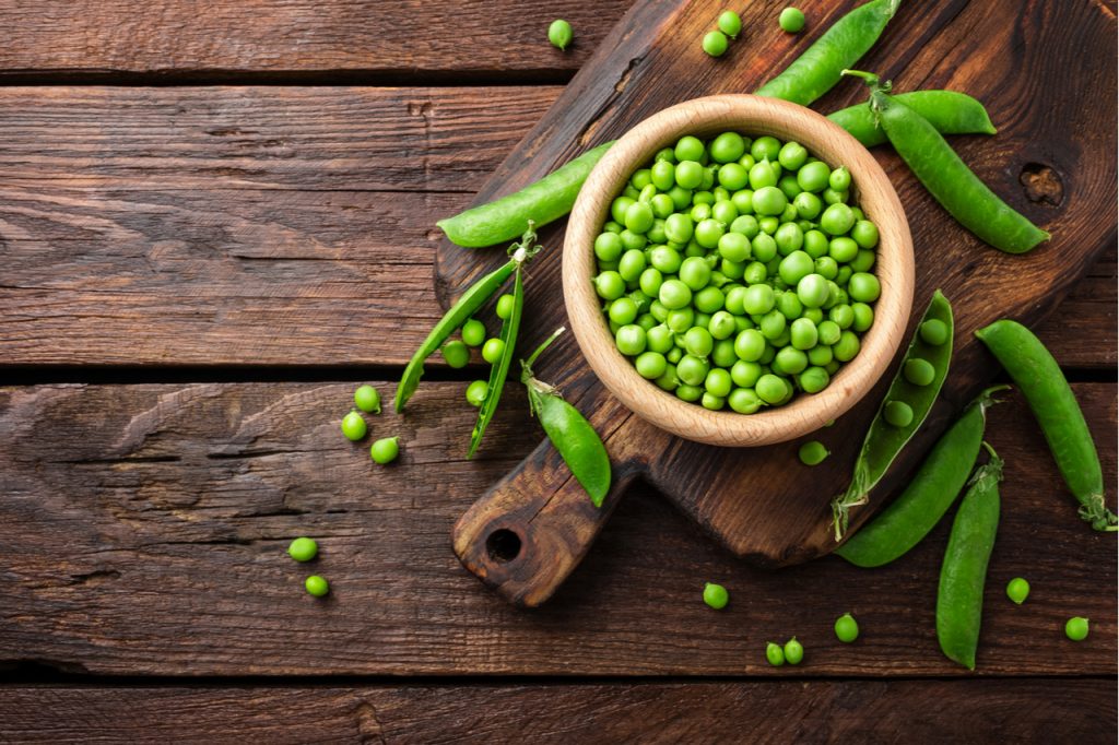 Green peas in a wooden bowl on a wooden chopping board.