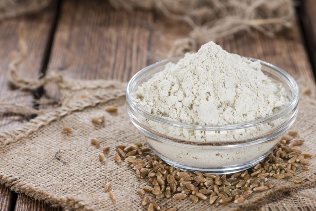 Spelt flour in a clear bowl on top of spelt seeds on a wooden table.