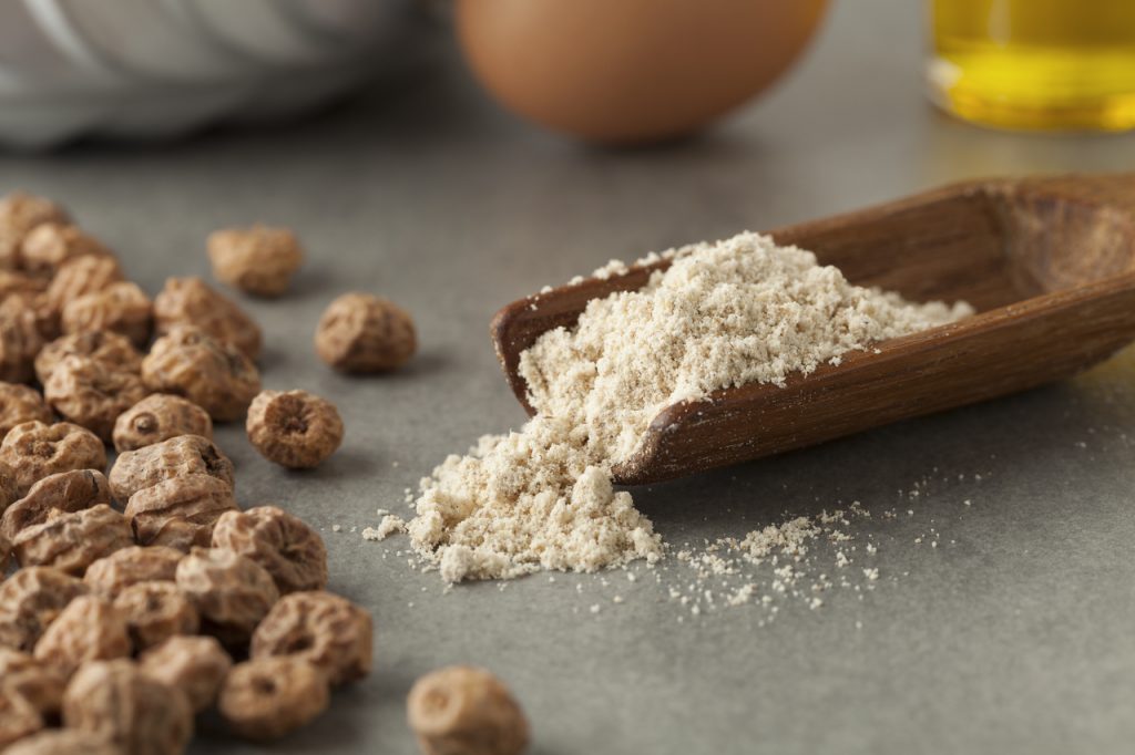 Tiger nut flour in a measuring spoon with scattered shelled tiger nuts on grey table.