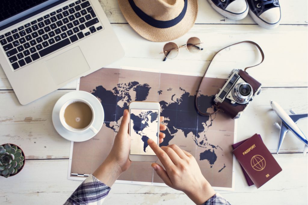 An image of a laptop, cup of coffee, sun glasses, pair of shoes, a hat, a camera, a model plane, 2 passport, a phone and a world map.