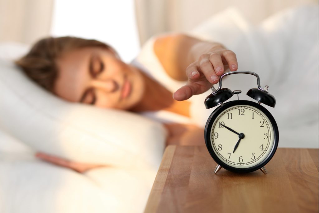 Woman stretching arm to alarm clock after waking up in the morning. What is your morning ritual?