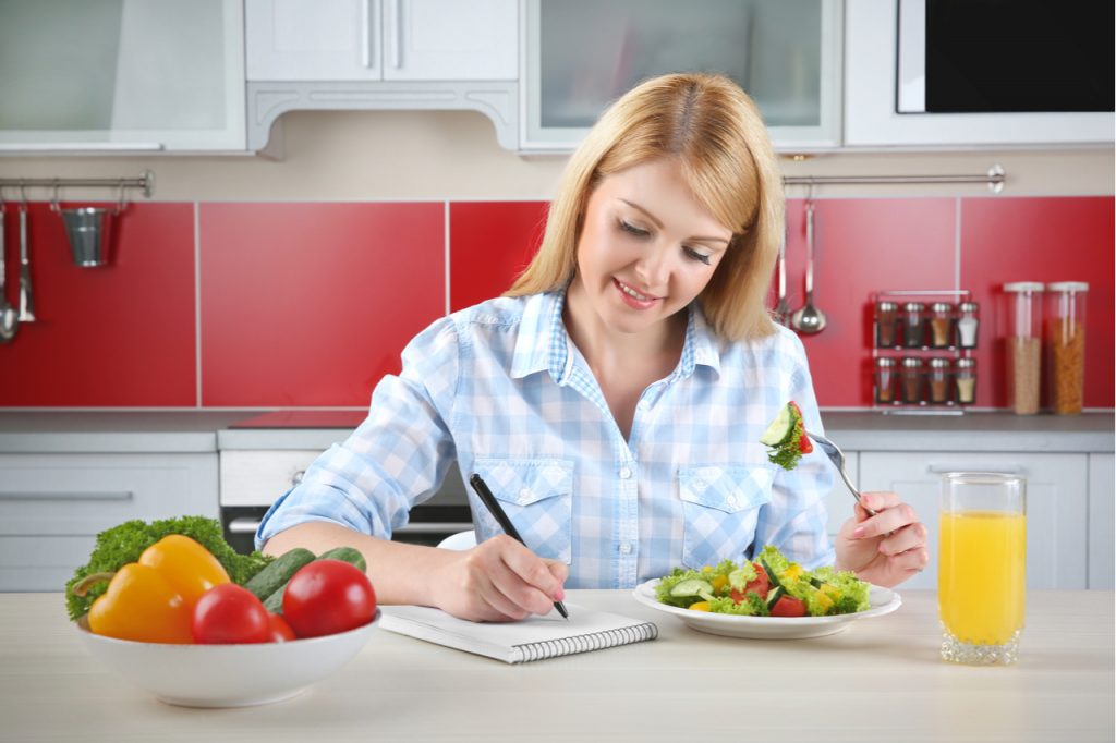 Woman eating salad while writing on food journal. Keeping track of your intake can help you if you are plateaued in your weight loss journey.