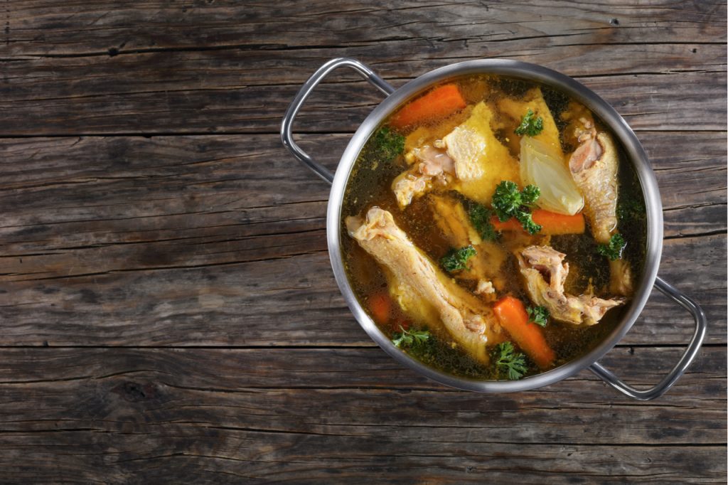 Chicken broth with pieces of rooster meat on bone and vegetables in a metal casserole on dark wooden table,