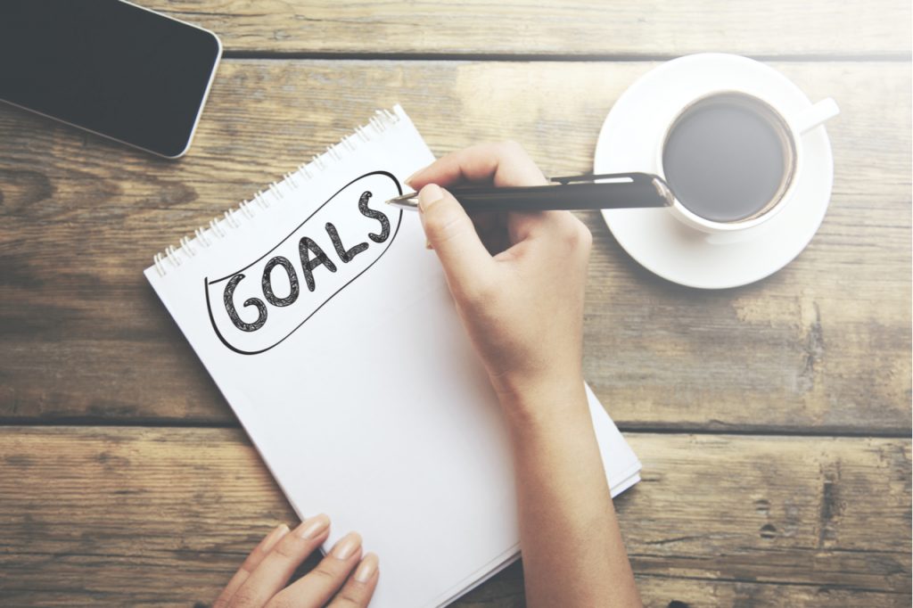 Someone writing goals on a notepad. A cup of coffee and cellphone next to the notepad on a wooden table.