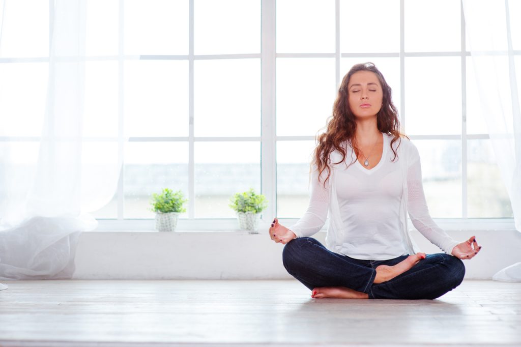 A woman meditating at home next to her window.
