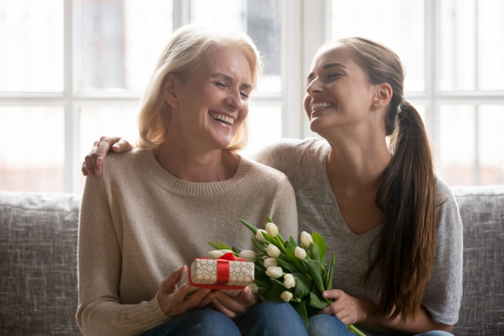 Mother and daughter sitting on the couch looking happy together. The daughter gave her mom a present and some flowers which looks to be a good mother's day gift idea.