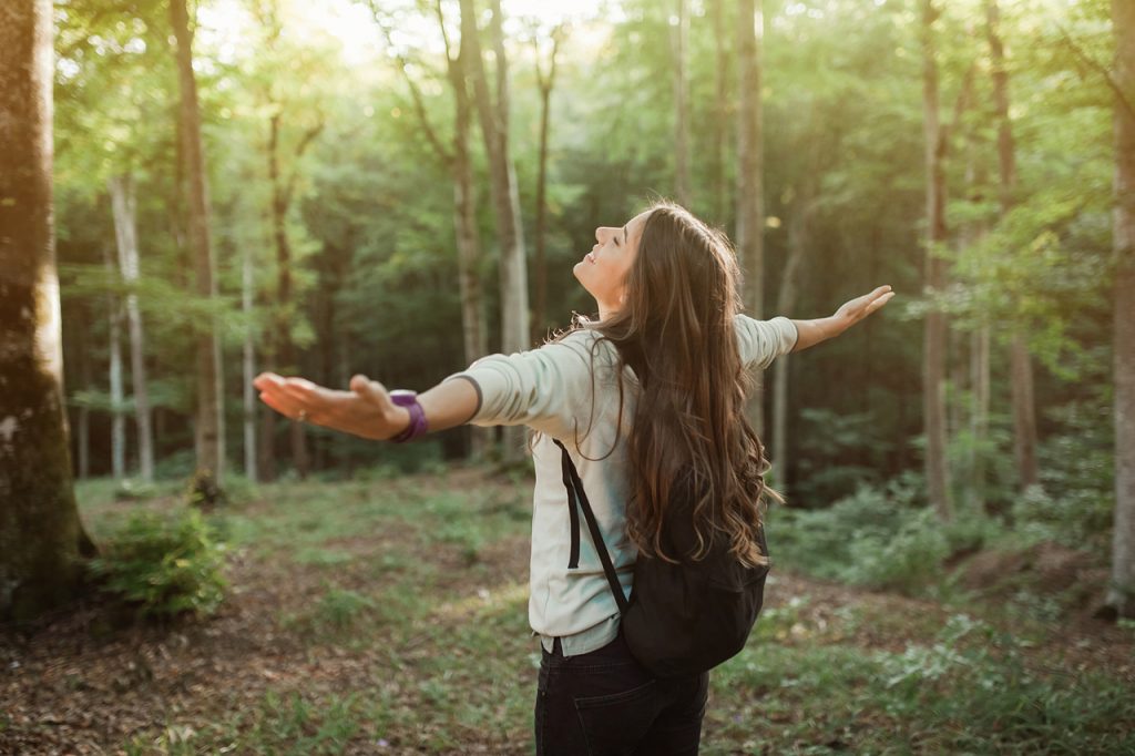 Woman emgracing the sun and nature with backpack in the forest on sunset light in the autumn season, looking up, exploring the nature.