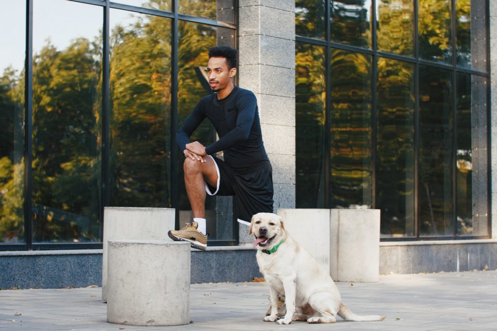 Young black man stretching after his training in city center with his white labrador dog beside him in the morning