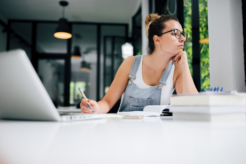 A woman sitting in front of her laptop holding her stylus while looking somewhere else.