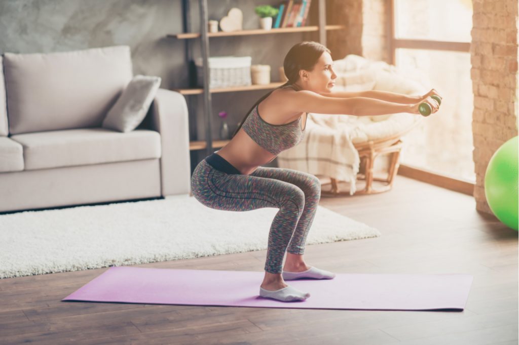 Woman doing deep squats workouts with dumbbells in outstretched arms.