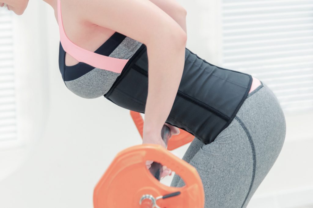 A girl in a corset and gray leggings with a barbell in the gym.