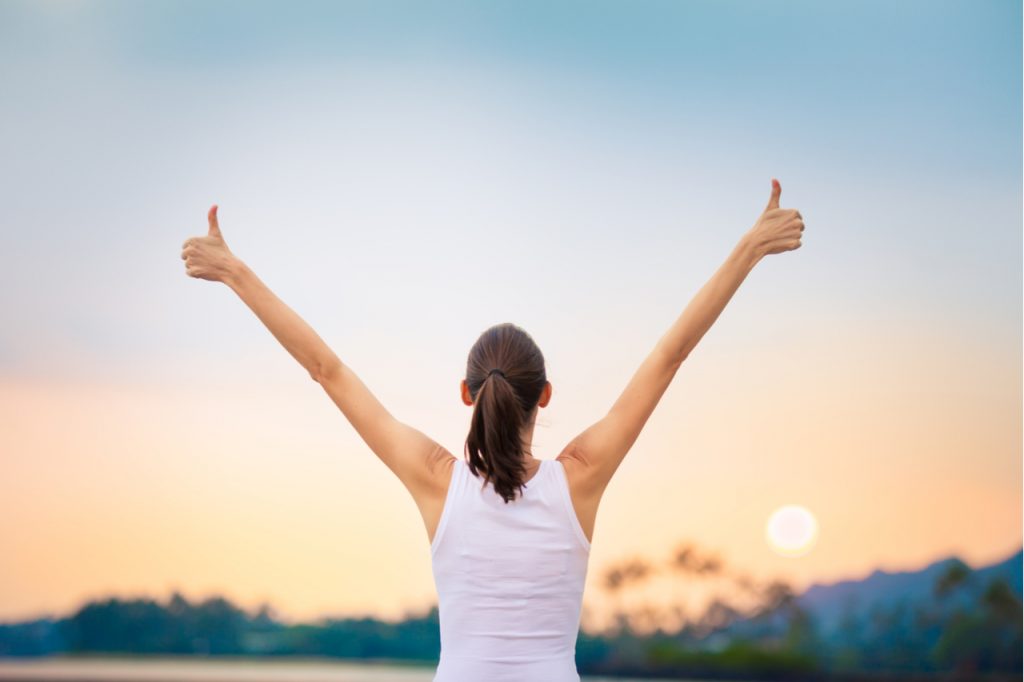Woman facing the sunset with both hands raised, showing a winning attitude.