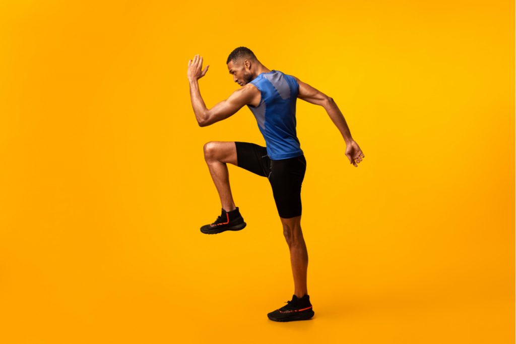 Energetic afro man doing fitness exercise, lifting leg and waving arm, free space