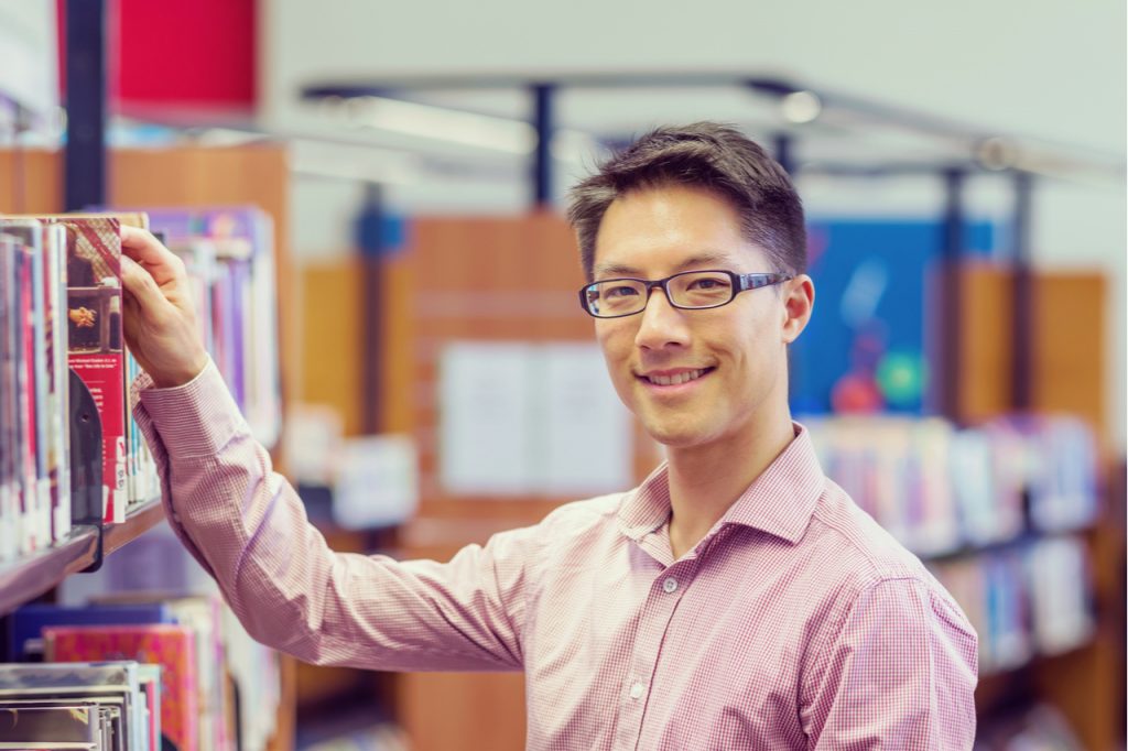 Intelligent looking man wearing glasses holding onto a book in a library.