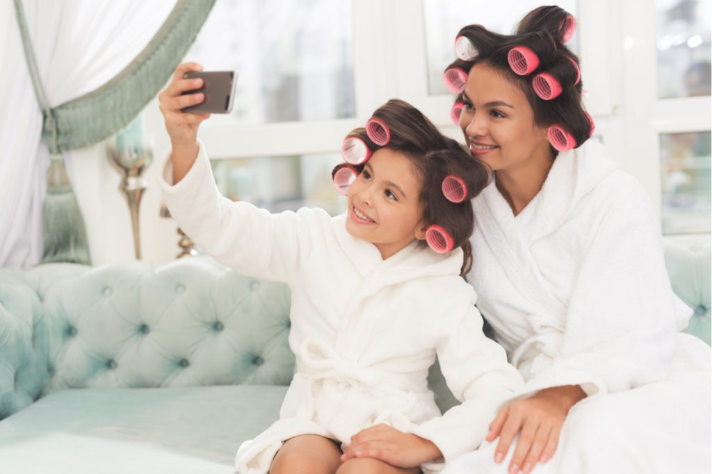 Mother and daughter in white bathrobes are sitting on the couch taking selfie.