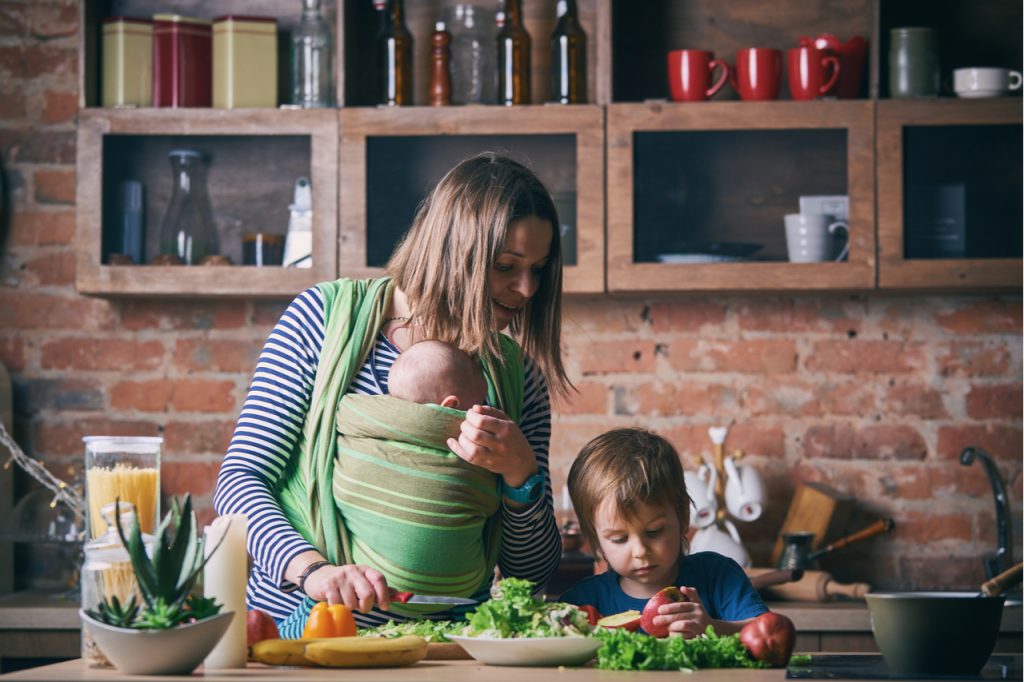 Mom and adorable boy and baby in sling cooking together in a sunny kitchen.