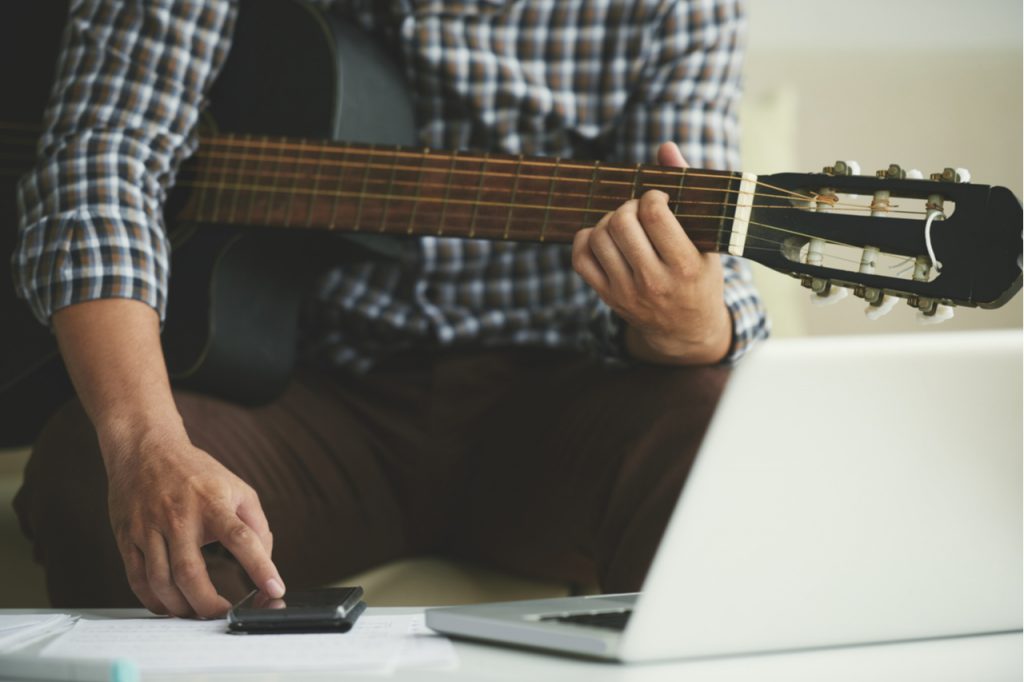Man writing notes in his smartphone when playing guitar at home.