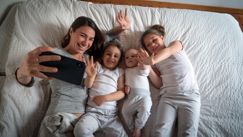  shot of happy mother with her kids are making a selfie or video call to father or relatives in a bed.