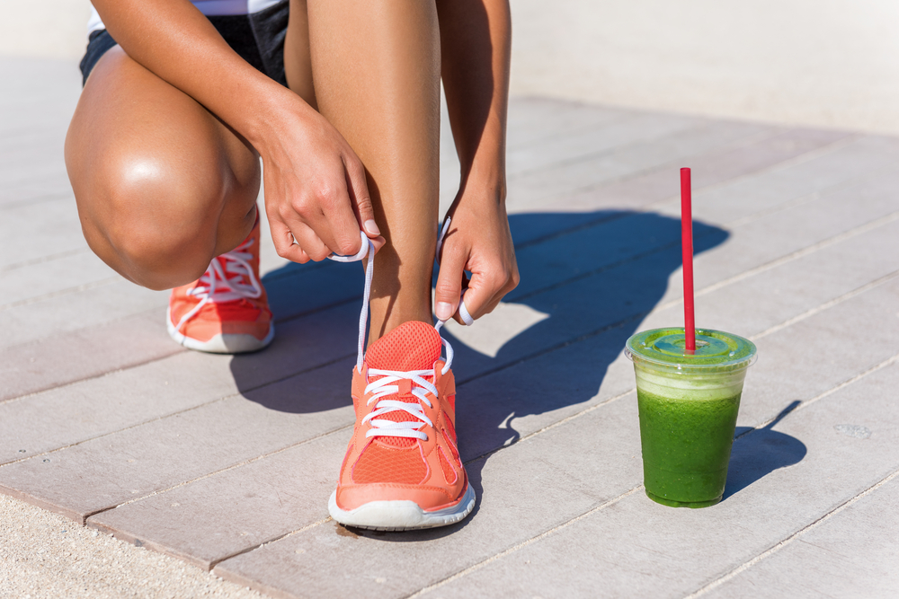 Running woman athlete runner getting ready for beach morning run by tying shoe laces of running shoes with green vegetable smoothie breakfast.