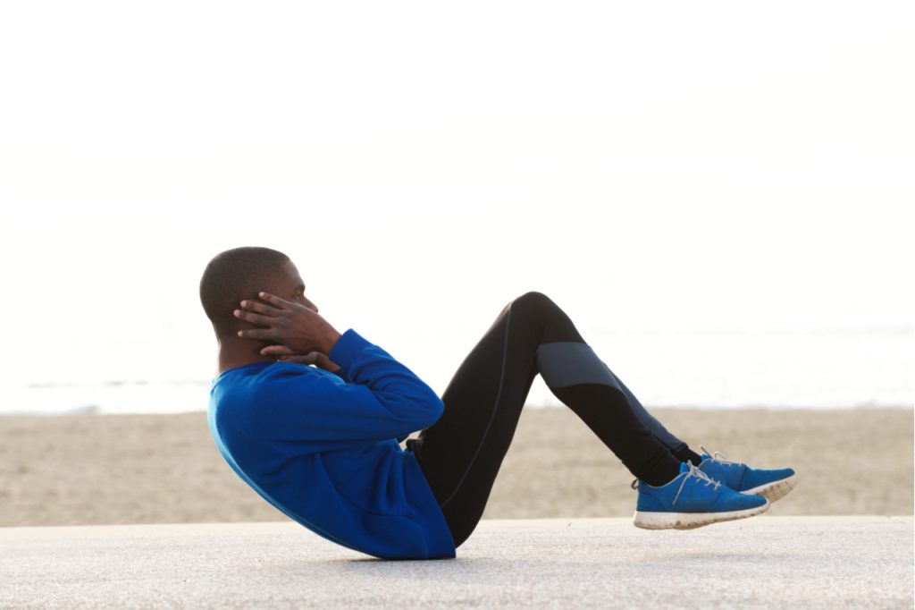 Side portrait of a young Afro man exercising on the beach doing sit ups.