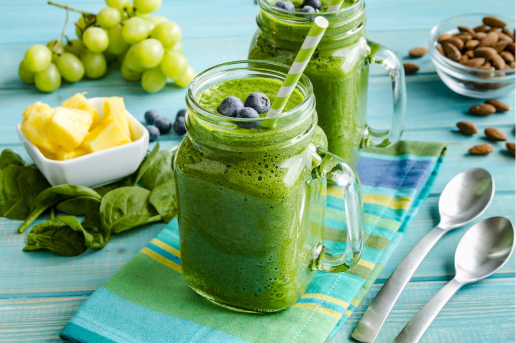 Mason jar mugs filled with green spinach and kale health smoothie with green swirled straw sitting with blue striped napkin and spoons.