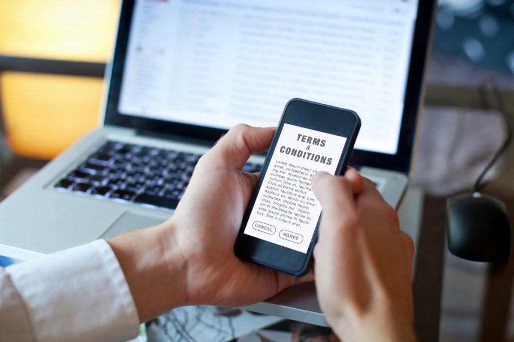 A man reading terms and conditions on his phone while sitting in front of his laptop.