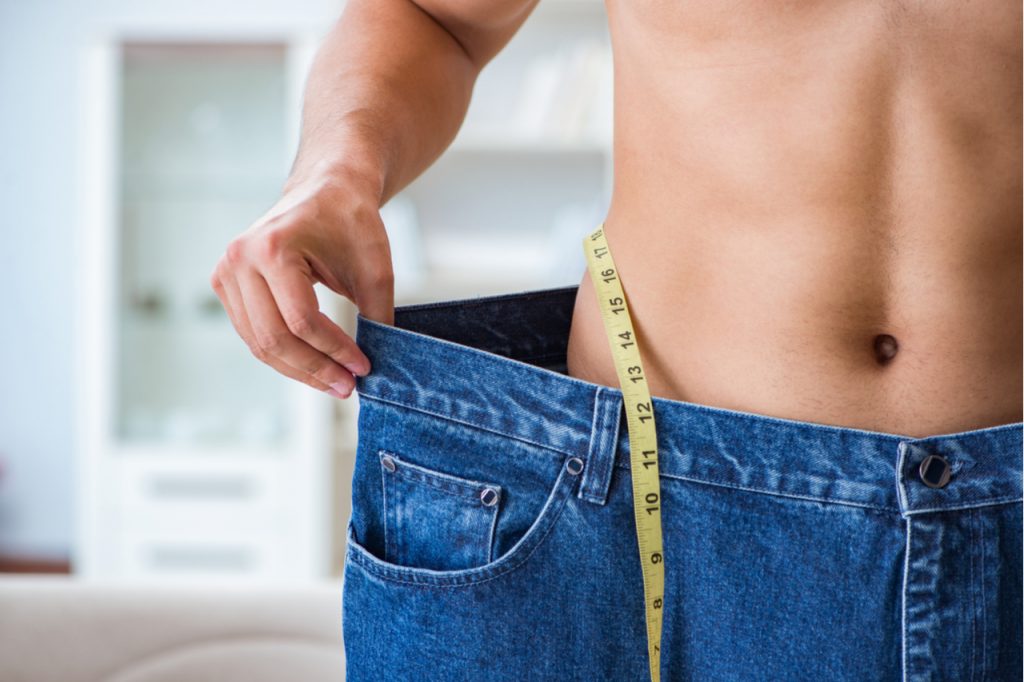 A man in oversized pants with a measuring tape, showing his weight loss success. 