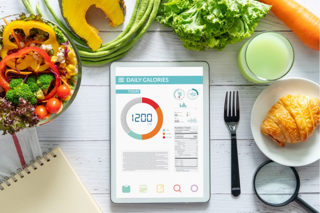 App on a tablet showing daily calories, next to some vegetables and a croissant on a white table.