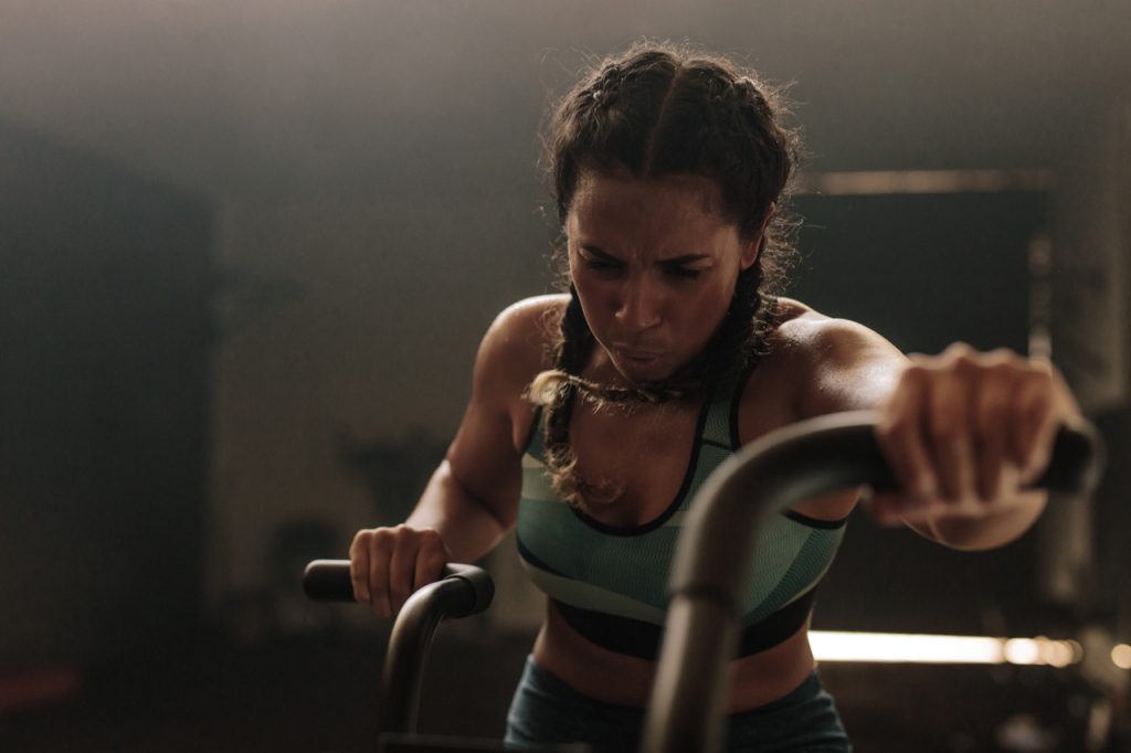 An African American woman working out on a stationary bike in a gym, thinking of her fitness motivation.