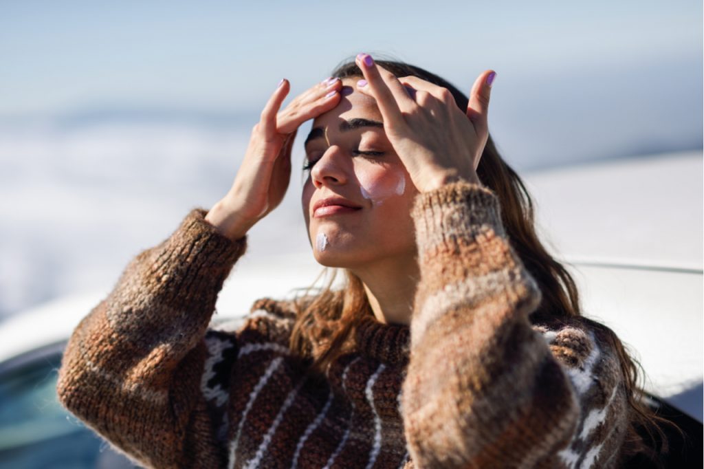 A woman on a sweater applying sun screen on her face for sun protection during winter months.