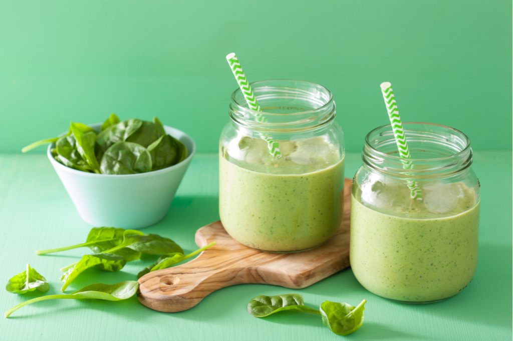 coconut green smoothie in 2 mason jars and a small bowl of spinach leaves on the side to relieve belly bloat.