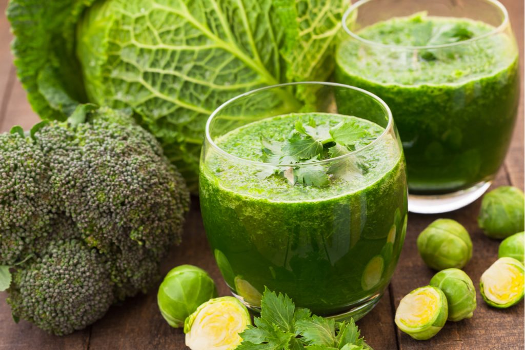 Two glasses of green smoothies with fresh broccoli, celery and Brussels sprouts surrounding the glasses. 