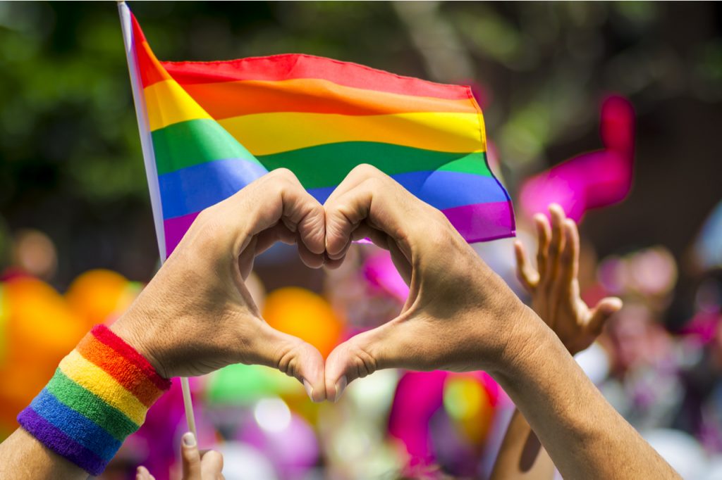 Hands forming a heart supporting pride and celebrating pride month with a rainbow flag in the background.