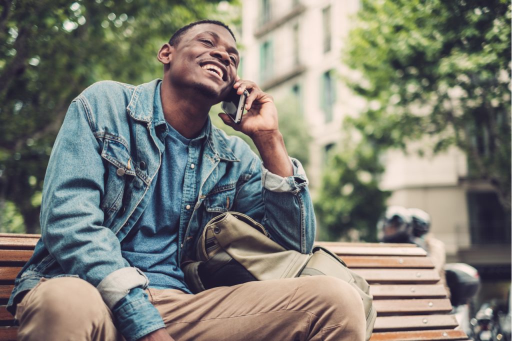 A man sitting on a bench at a park while happily talking to a friend on the phone.