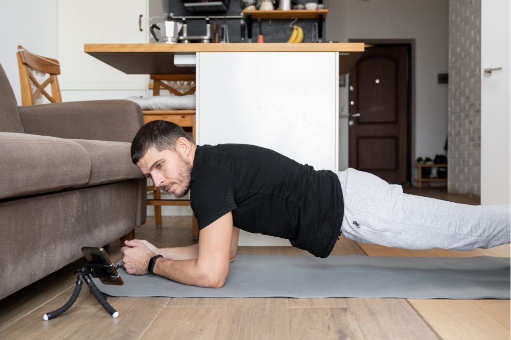 A man doing planking in the living room while watching a virtual class on his phone. 