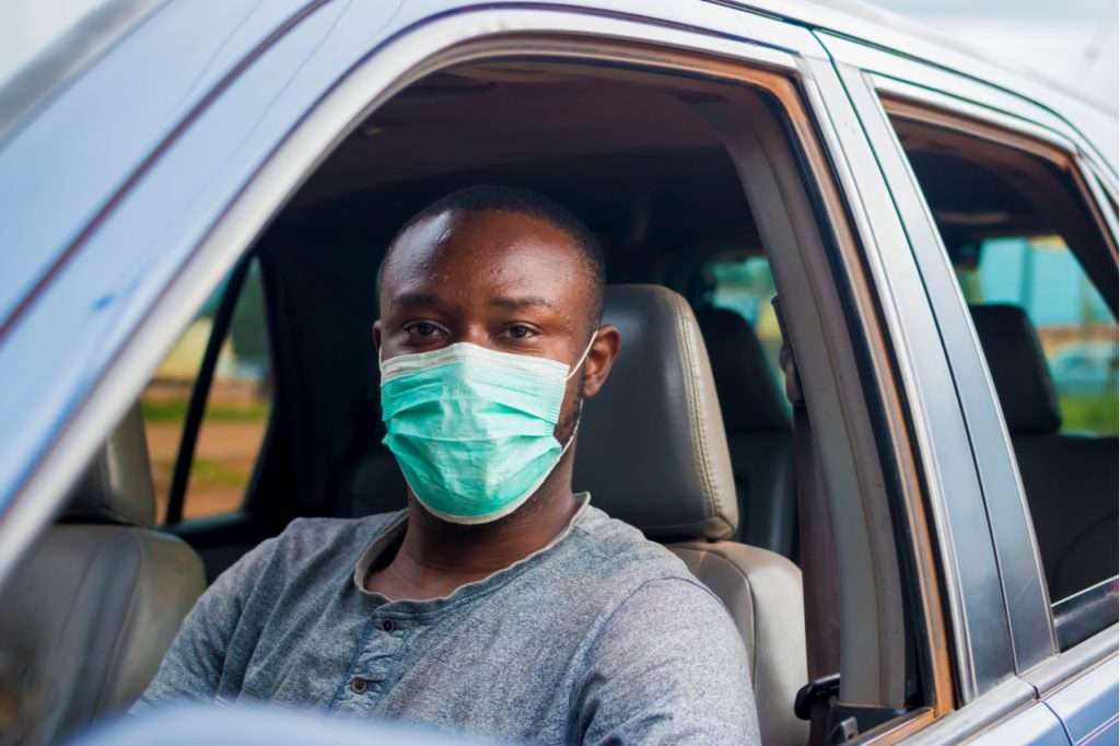 A man wearing a face mask for protection sitting at the driver seat of a car.