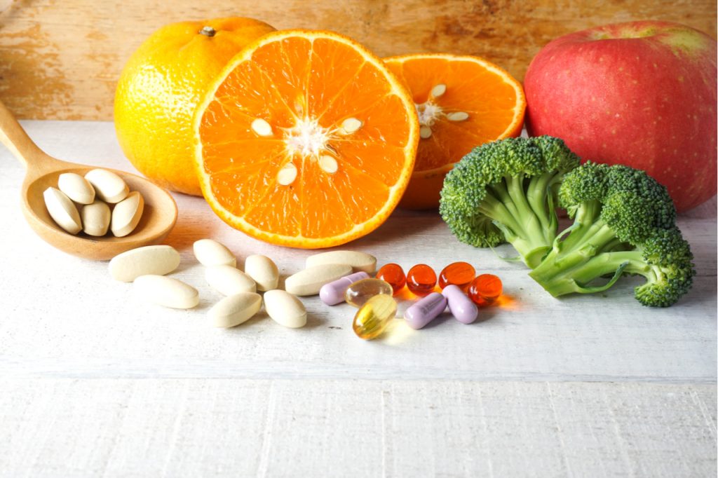 Multivitamins in capsules and gel tablets with broccoli, apple and oranges on the side.