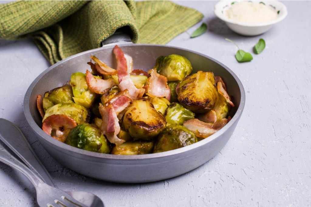 Oven Roasted Parmesan Brussels Sprouts With Bacon in small pan with a spoon and fork on the side and a table napkin. 