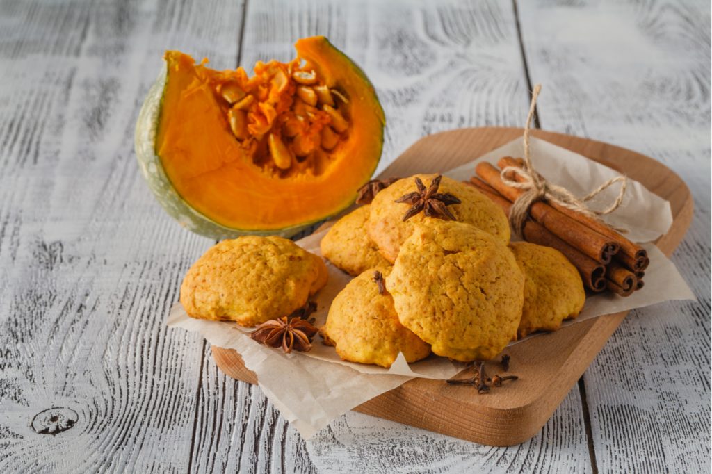 Spiced pumpkin cookies on a wooden board with some sticks of cinnamon and a sliced pumpkin. 